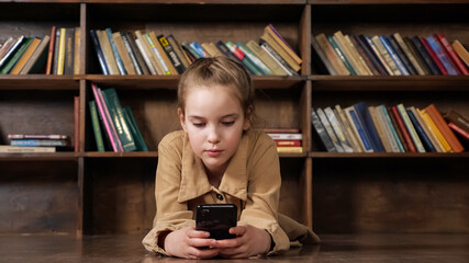 Girl junior student in brown jacket plays online game on black smartphone sitting against wooden...