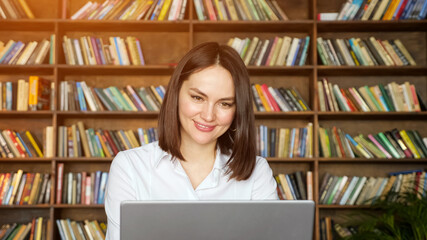 Woman freelancer with short hair talks online on near grey laptop against large bookshelves in local library, sunlight