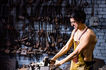 Fototapeta na wymiar Authentic blacksmith man forges a metal product in dark indoors studio
