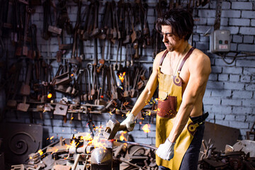 Obraz na płótnie Canvas Authentic blacksmith man forges a metal product in dark indoors studio