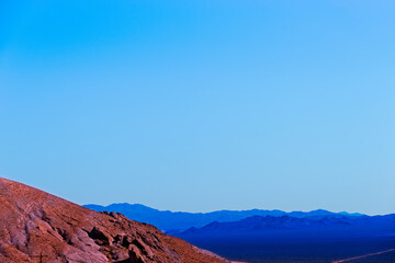 Barren reddish barren mountainside with purple and blue mountain ranges beyond under a hazy blue sky.