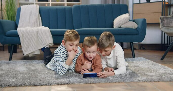 Lovely smiling happy different ages three boys lying on the floor and using smartphone to watch cartoon