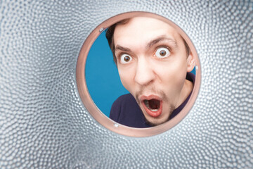 Man looking inside cup or rounded metal tube. Close-up shot of shocked young man with round eyes and open mouth.