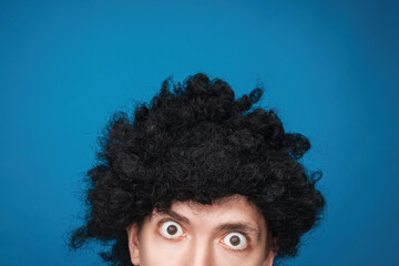 Funny portrait of a man with happy emotion on his face in the studio on blue background. Man wearing wig. Black hair