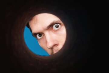 Man's eyes looking inside rounded tube. Empty place for a text or object. Close-up shot of shocked young man with round eyes