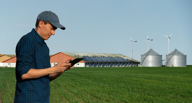 Farmer With Tablet Computer On A Background Of Modern Dairy Farm Using Renewable Energy, Solar Panels And Wind Turbines