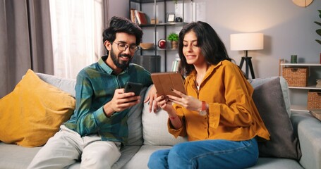 Portrait of Indian cheerful young family couple sitting on couch in apartment using digital tech gadgets. Pretty wife browsing online on tablet while her husband using smartphone, marriage concept