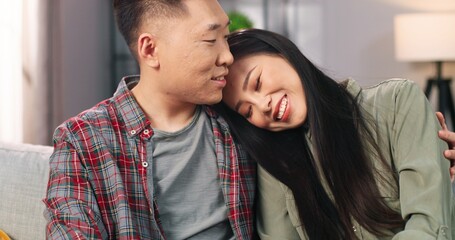 Close up of happy Asian young beautiful woman speaking with husband while sitting on couch in cozy room in apartment. Cheerful married couple in love spending time together in positive mood at home
