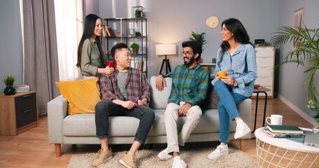 Fototapeta na wymiar Joyful young mixed-races family couples sitting on sofa in modern living room and laughing in good mood. Asian and Hindu men and women at home drinking coffee or tea and speaking, friends concept