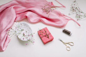 Overhead view of essential beauty items, top view of pink silk fabric, natural soap, flowers and decorative scissors, flat lay isolated on white background