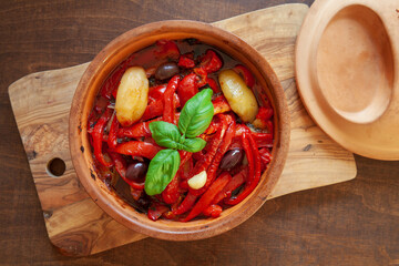 Roasted bell peppers with potatoes, olives, garlic, basil leaves and tomato sauce in clay pot. Italian cuisine peperonata dish.
