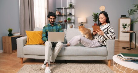 Cheerful young mixed-race wife and husband couple resting on couch in modern living room holding cat pet browsing on digital gadgets, Hindu man typing on laptop while Asian woman working on tablet