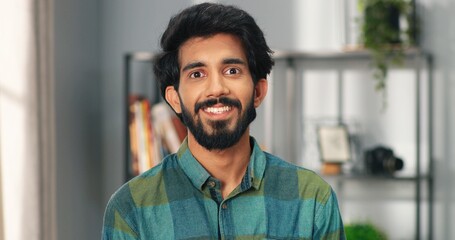 Fototapeta premium Close up portrait of happy joyful Hindu handsome man stands in room in house, looking at camera and smiling. Positive emotions. Young millennial guy at home with smile on face alone, good mood