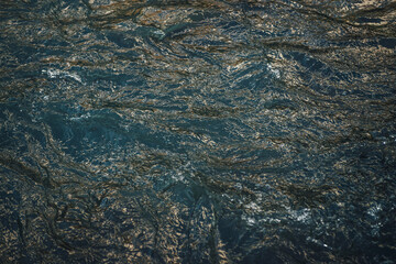 Atmospheric nature background of golden water of mountain lake in sunrise or sunset. Scenic fragment of water surface with gold sunshine reflection. Sunlight reflected in golden blue water stream.