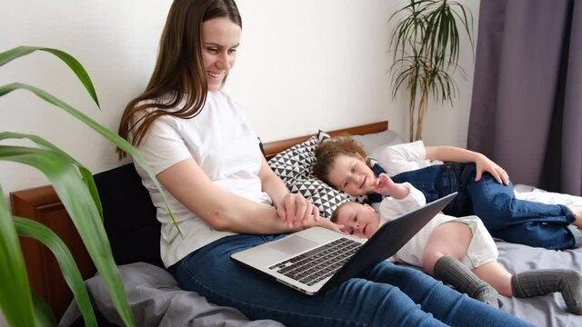 Relaxed beautiful young mom with cute preschool daughter and baby infant rest on comfy bed spend weekend laughing watching family cartoons on laptop, enjoy free time at home. Modern tech usage concept