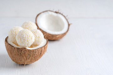 Fototapeta na wymiar Coconut truffles or homemade organic energy balls with a filling of sweetened shredded coconut and curd cheese served in halved fresh coconut on white wooden table. Image with copy space, horizontal
