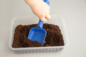 A child's hand pours peat with a shovel into a container. Part 1 of step by step instructions