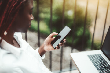 View with a shallow depth of field and selective focus on the smartphone in a hand of a young black female freelancer with braided hair, sitting on the balcony with her laptop in front of her