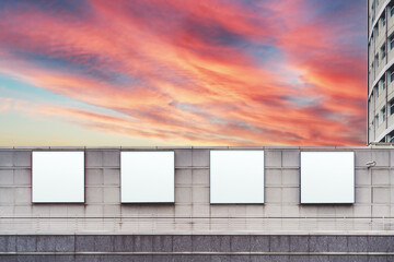 Obraz premium Four empty white square billboard mockups on the facade of a modern shopping mall or a business center as advertising banners placeholders, with a beautiful sunset sky in a defocused background