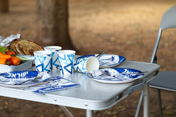 Folding furniture for a picnic in the forest. The table covered with disposable dishes with the symbols of Israel (with the inscription Israel in Hebrew). Yom Haatzmaut (Israel Independence Day)