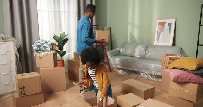 Young African American joyful cute family couple unpacking their stuff spending time together in new apartment on moving day. Wife and husband move and open carton boxes in own home. Tenant concept