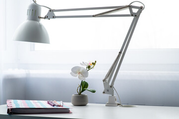 A simple white desk in a homeworker. There is a white lamp on the table. There is a large notebook on the table. The pens are on the notebook.
