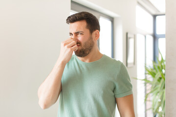 handsome man feeling disgusted, holding nose to avoid smelling a foul and unpleasant stench