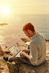 A young man uses his laptop to telecommute remotely with the sea in the background