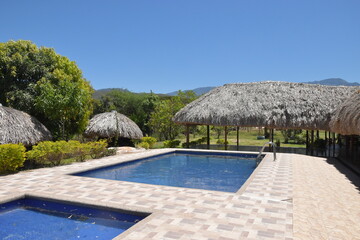 Piscina con chozas al rededor, cielo azul y arboles entre jadín