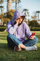 Bored teenager wearing a purple hoodie is using a mobile phone in the park. She is sitting in the...