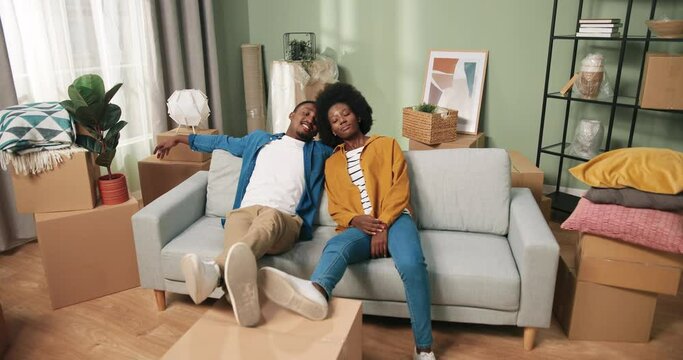 Camera approaching joyful young family wife and husband sitting on couch tired after unpacking boxes and decorating new apartment. African American couple resting after relocation and settle in