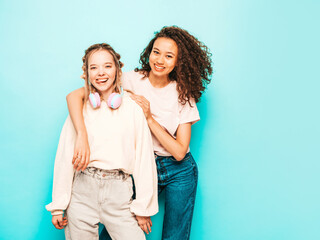 Two young beautiful smiling international hipster female in trendy summer clothes. Sexy carefree women posing near blue wall in studio. Positive models having fun. Concept of friendship