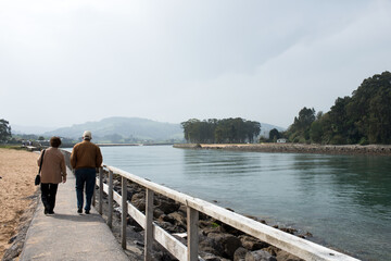 Aged couple 70-80 walking in Rodiles seen from their back. Wooden path next to the river with Puntal in the background. Asturias.