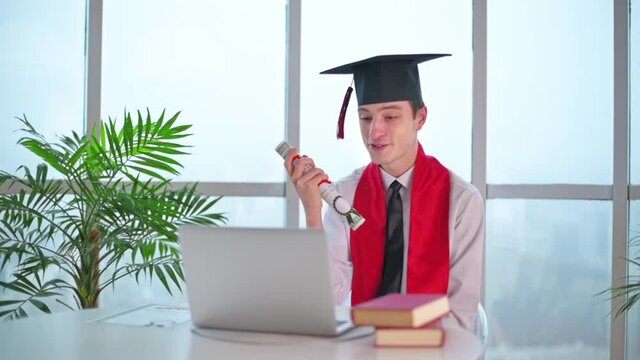 Young man with graduation diploma talking on the laptop at home. Graduating online in virtual ceremony