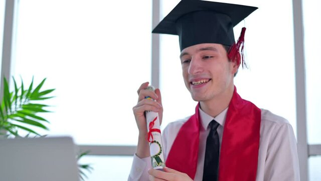 Young man with graduation diploma talking towards the camera. Graduating online in virtual ceremony