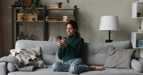 Happy young beautiful woman sitting on comfortable sofa, wearing wired earphones, listening music in mobile audio library application, communicating distantly, enjoying weekend in living room.