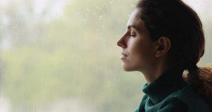 Side close up head shot view mindful young woman breathing fresh air with closed eyes near window with rainy weather outside, doing yoga exercises, meditating alone indoors, enjoying peaceful time.