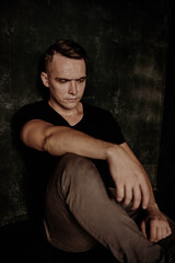 The man sits next to the wall looking forward, the room is dim.