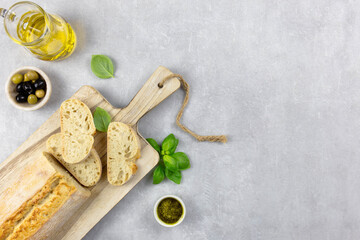 Fresh italian ciabatta bread with herbs, olive oil, black and green olives, basil leaves and pesto sauce on light concrete background. Top view. Copy space.