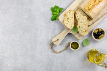 Fresh italian ciabatta bread with herbs, olive oil, black and green olives, basil leaves and pesto sauce on light concrete background. Top view. Copy space.