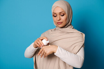 Portrait of beautiful woman in hijab squeezing cream on hand, isolated on blue background with space for text