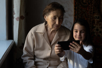 Great-grandmother sits with great-granddaughter and looks into the smartphone. Grandmother and child take a selfie on a smartphone. Grandmother with her granddaughter look into the phone.