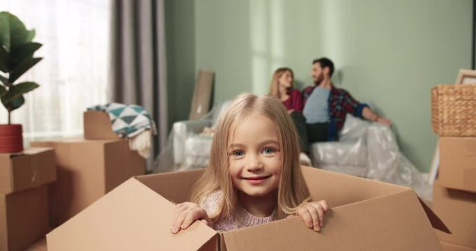 Close up of Caucasian little cute girl child sitting in big carton box in good mood having fun playing and smiling to camera while parents sitting on sofa behind. Unpacking on relocation day Settle in