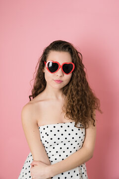 Beautiful young woman with curly brown hair , heart shape sunglasses and cute polka dot dress on bubblegum pink background