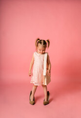 little girl in a festive dress and her mother's shoes and with a bag looks down on a pink background with a place for text