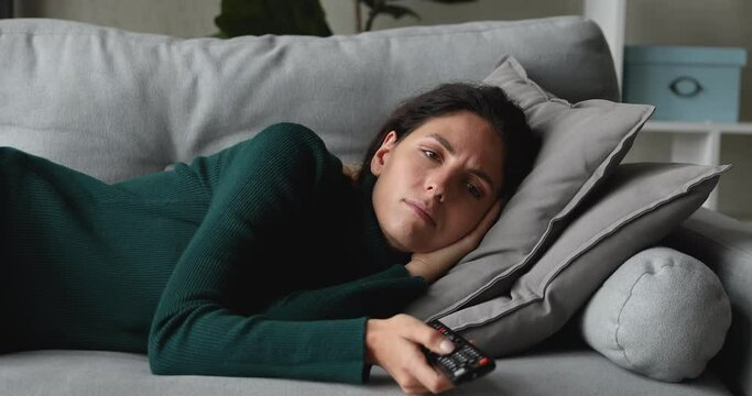 Unhappy young woman lying on sofa, switching programs with remote controller, searching for interesting TV show series funny movie, feeling bored spending free weekend leisure time alone at home.