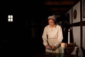 Eine Frau steht im alten Haus und putzt  mit dem Lappen beim Kerzenlicht das Geschirr ab. Sie ist angezogen wie eine Bäuerin mit dem Kopftuch, einem alten Kleid und einer Schürze. 