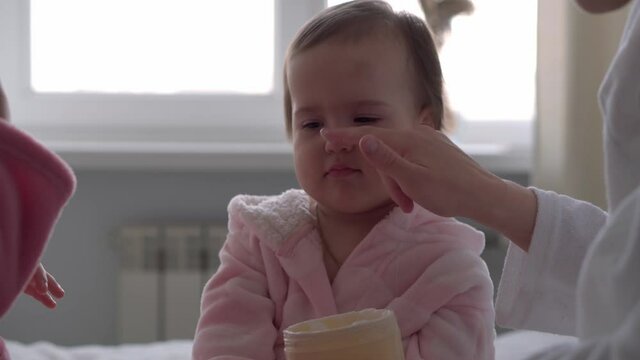 Authentic cute young woman applying face mask of baby girl. mom with 2 children daughters different ages in bathrobes after bath on white bed oposite window. Spa, motherhood, childhood, family concept