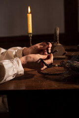 Eine Bäuerin sitzt am Tisch beim Kerzenlicht  in einem alten Haus und macht aus den Pferdehaaren ein Armband. Überall liegt Schmuck, antik Bügelleisen und es brennen . Kerzen.  Sie hat ein Kopftuch an