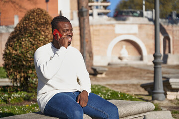Fototapeta premium Happy black man talking on smart phone sitting in a park on a sunny day. 